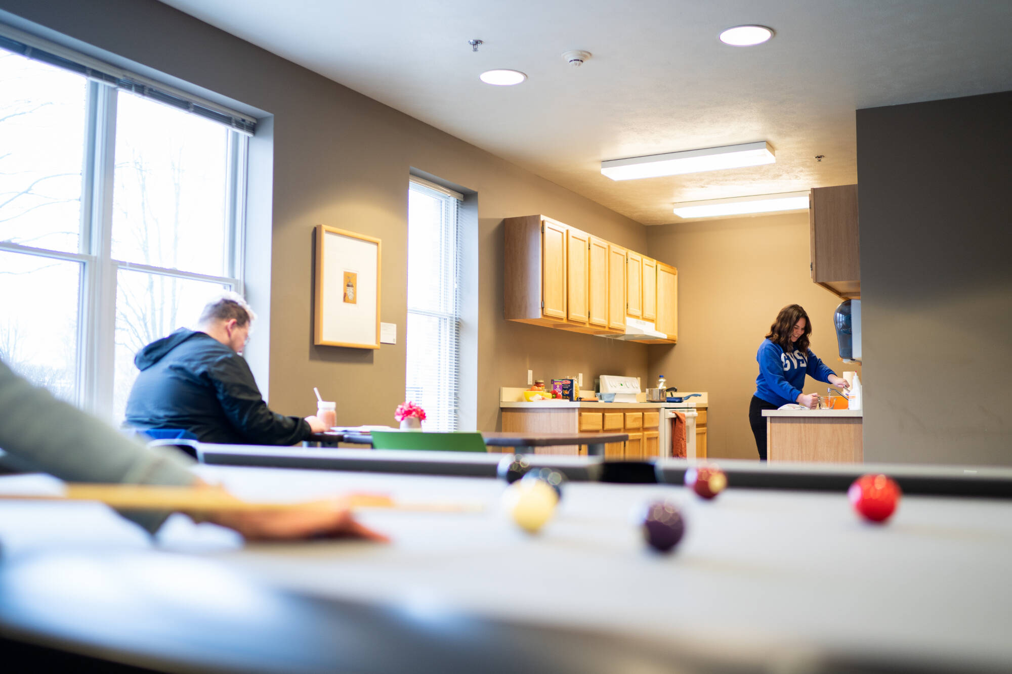 Winter Hall community area with a kitchen and living area with a pool table. One student studies at a table while another cooks in the kitchenette.